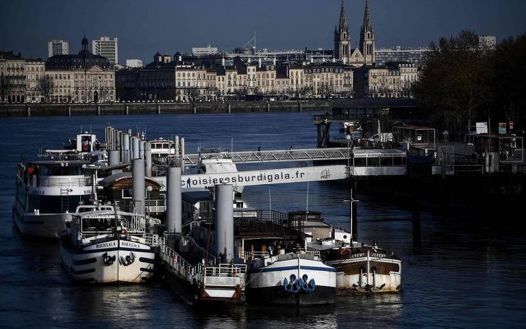 Vários barcos de passeio estão atracados em um cais no rio, com uma passarela que exibe o endereço 'croisieresburdigala.fr'. Ao fundo, há prédios históricos e duas torres de igreja sob céu claro.