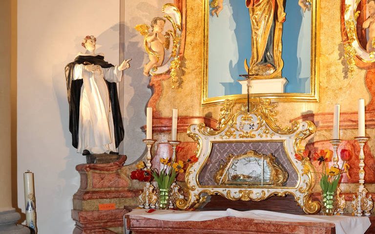Altar em igreja com estátua de santo à esquerda e imagem de Nossa Senhora ao centro, cercado por velas acesas e vasos de flores. Cordão vermelho delimita o espaço do altar.
