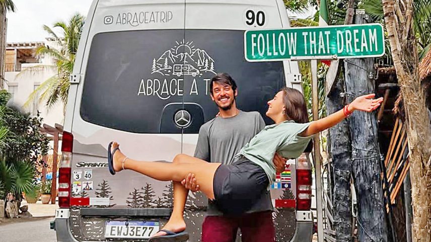 Homem segura mulher nos braços em rua com palmeiras. Van branca ao fundo exibe logo e texto 'Embrace a T'. Placa verde com texto 'Follow That Dream' está visível.