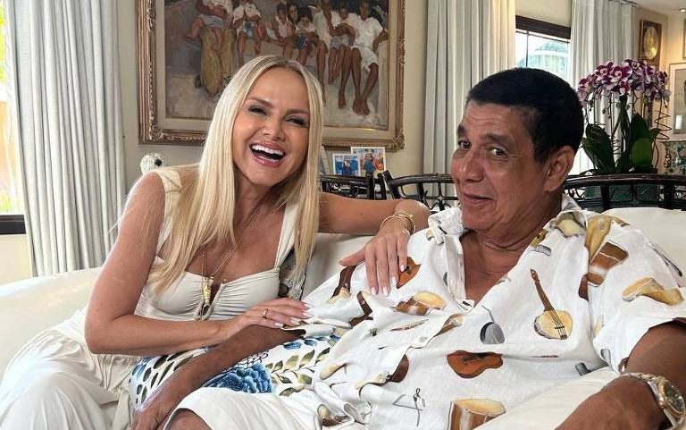 Homem e mulher sentados lado a lado em sofá branco. Mulher loira veste roupa clara e sorri, homem usa camisa branca estampada e shorts, com expressão neutra. Ao fundo, quadro grande e cortinas claras.