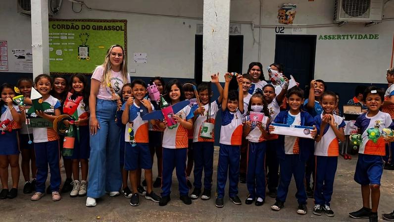 Vinte crianças em uniforme escolar azul, branco e laranja posam com uma professora loira de calça jeans e camiseta branca em pátio de escola. Crianças seguram brinquedos e sorriem diante de mural verde e portas com placas.