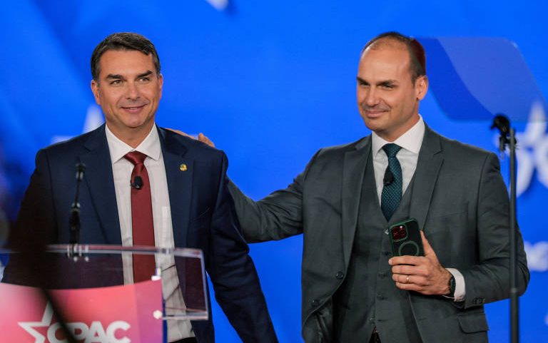 Dois homens de terno estão lado a lado em palco com fundo azul. Um deles está atrás de um púlpito com logo CPAC e o outro toca seu ombro direito segurando um celular.
