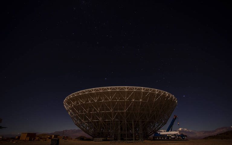 Radiotelescópio grande em formato de prato parabólico instalado em área desértica. Céu escuro com estrelas visíveis ao fundo e montanhas ao longe. Equipamento de construção e guindaste próximos à estrutura.