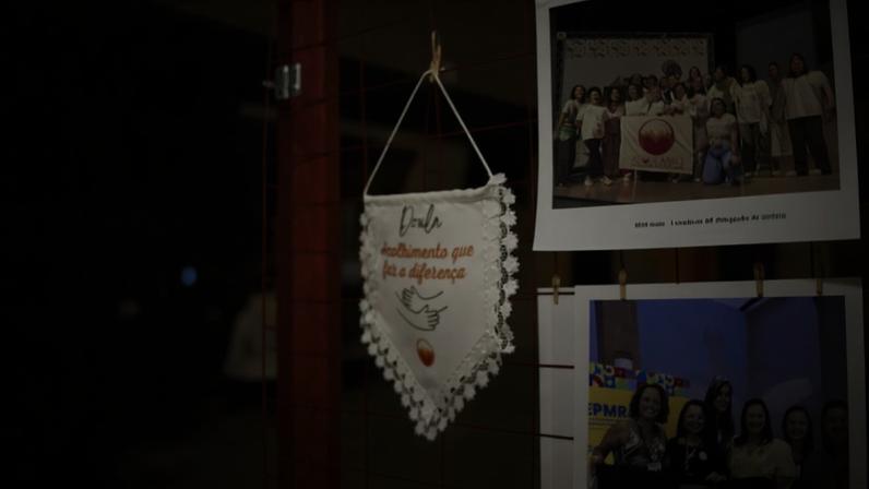 Estandarte branco com borda de renda pendurado em grade vermelha, com texto "Doula acolhimento que faz a diferença" e desenho de mãos se abraçando. Ao fundo, fotos desfocadas de grupos de pessoas em eventos.
