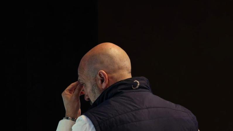 Homem calvo de costas, vestindo colete escuro e camisa branca, observa atentamente várias telas com gráficos e tabelas financeiras coloridas em ambiente de trabalho.