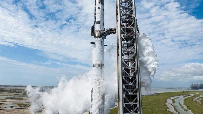 Foguete preto e branco posicionado verticalmente em plataforma de lançamento, com emissão visível de vapor branco saindo da base. Estrutura metálica e área de concreto ao redor são visíveis em vista aérea.
