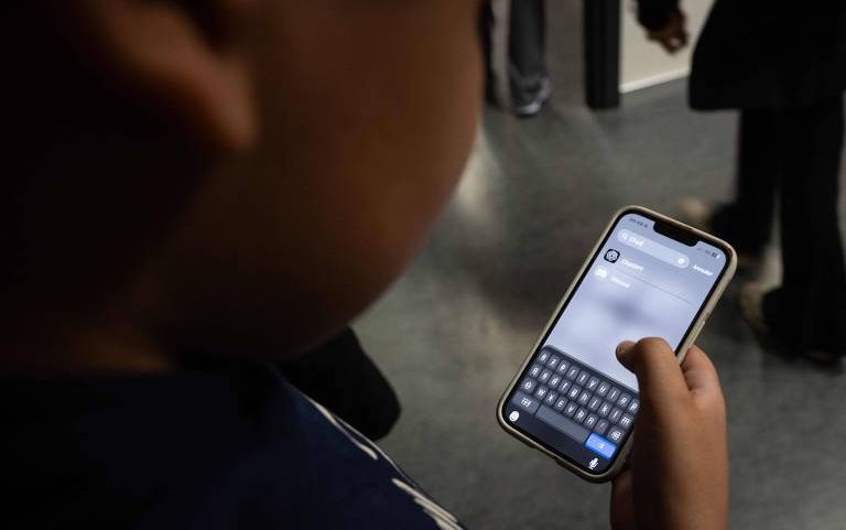 Pessoa segura smartphone com tela mostrando aplicativo de mensagens aberto e teclado virtual ativo. Fundo desfocado indica ambiente interno com outras pessoas em pé.