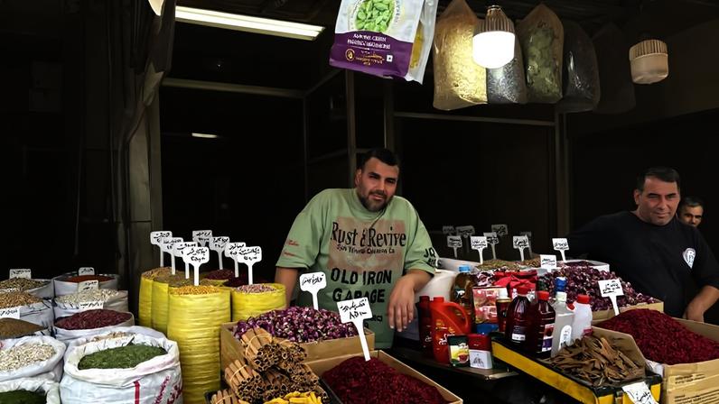 Homem atrás de balcão em barraca com sacos e caixas cheios de especiarias, grãos e ervas secas. Rótulos indicam tipos variados, como açafrão e hibisco. Ambiente interno de mercado com iluminação artificial.