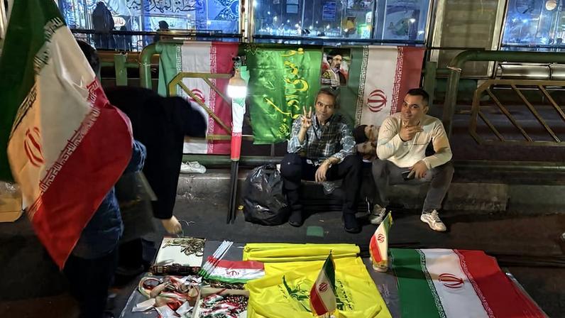 Três pessoas em ponto de ônibus à noite com bandeiras do Irã e acessórios à venda no chão. Duas pessoas sentadas atrás das bandeiras exibem sinais de paz com as mãos. Bandeiras penduradas no fundo e iluminação artificial no local.