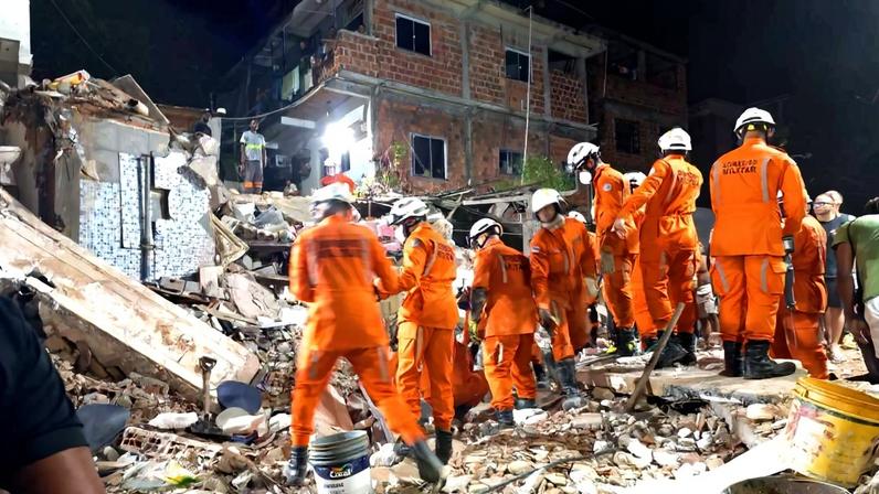 Dez socorristas com uniformes laranja e capacetes brancos trabalham entre escombros de um prédio parcialmente destruído durante a noite. Ao fundo, edifício de alvenaria com janelas iluminadas.