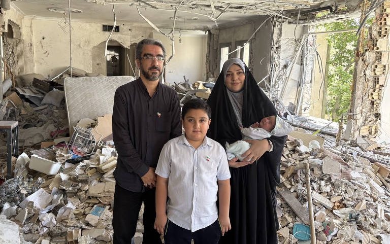 Homem, mulher com hijab segurando bebê e menino posam em sala destruída com paredes e teto danificados, entulho espalhado e janela aberta para área externa.