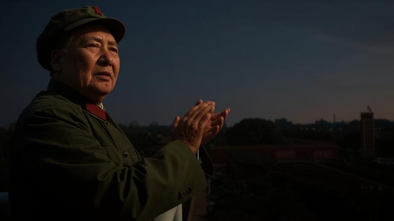 Homem em uniforme verde militar com boina, de perfil, aplaude olhando para multidão abaixo. Multidão densa se estende ao longo de uma avenida com árvores e edifícios ao fundo.