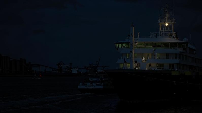 Navio grande atracado em cais de porto industrial, com rebocador próximo na água. Ao fundo, estruturas industriais e guindastes visíveis sob céu parcialmente nublado.