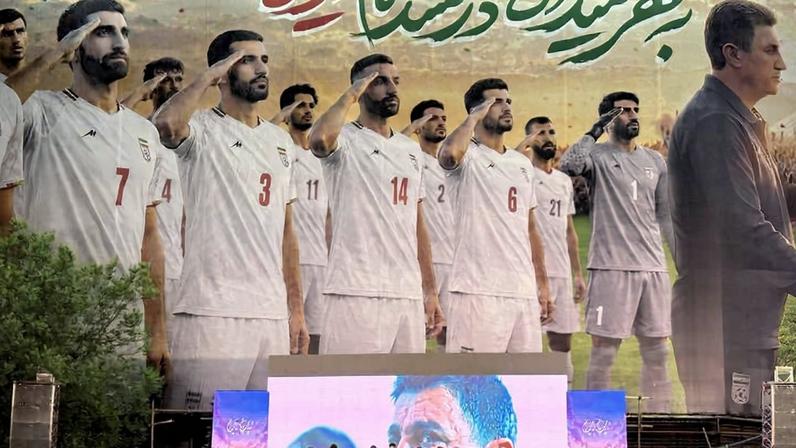 Equipe de futebol em palco diante de grande mural com jogadores em uniforme branco fazendo continência. Multidão com bandeiras e celulares erguidos observa o evento ao ar livre.