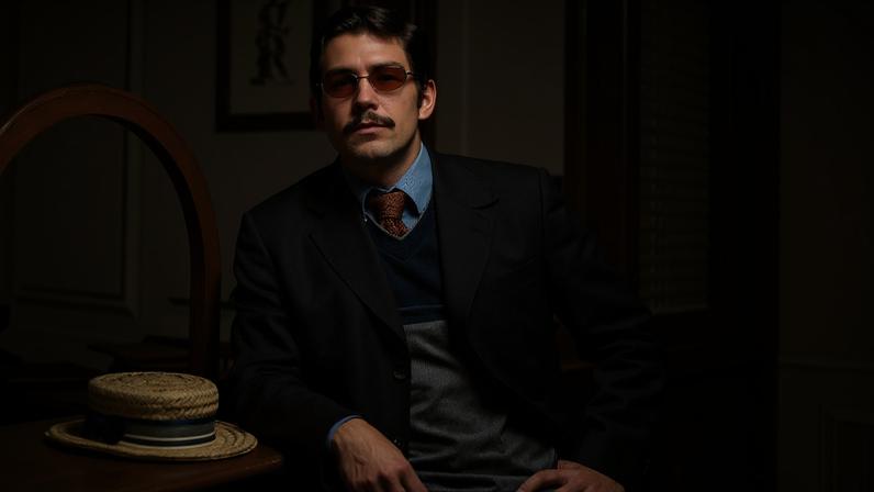 Homem de cabelo curto e bigode usa óculos escuros, terno escuro, suéter cinza e camisa azul com gravata marrom, encostado em balcão de madeira. Chapéu de palha com fita azul está sobre o balcão. Ambiente interno com porta e quadro ao fundo.