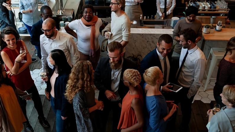 Grupo diversificado de pessoas em ambiente interno, algumas segurando copos e conversando em pequenos grupos. Ambiente com piso de madeira e balcão ao fundo com utensílios e bebidas. Pessoas vestem roupas casuais e formais, interagindo em clima descontraído.