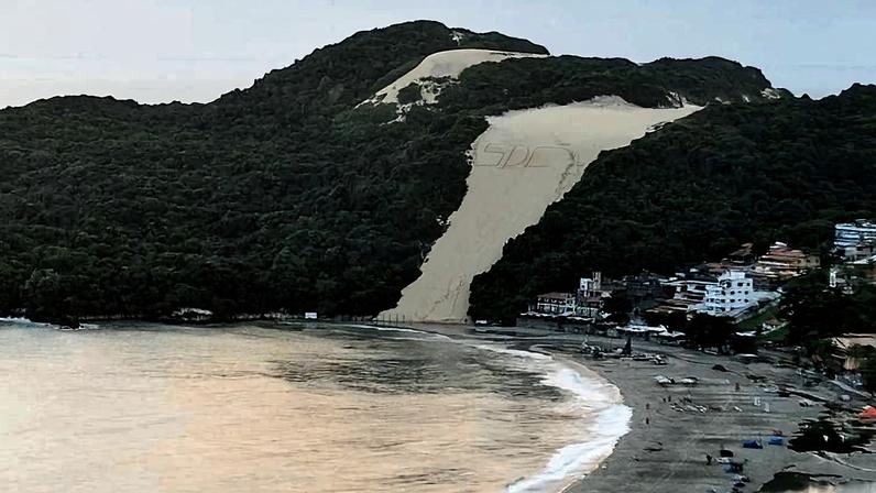 Duna de areia clara no Parque das Dunas em Natal, cercada por vegetação verde densa. Praia com faixa de areia e ondas suaves à direita da duna. Edificações residenciais e comerciais próximas à praia, com algumas pessoas e guarda-sóis visíveis.