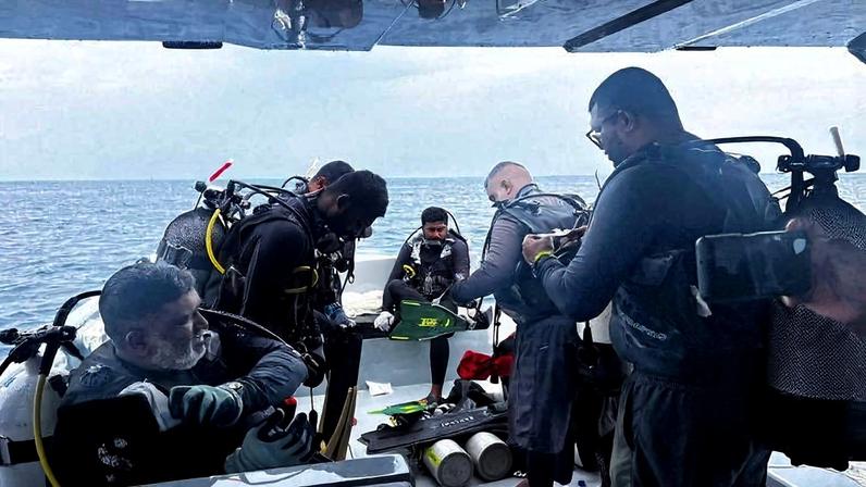 Seis mergulhadores equipados com cilindros de oxigênio e roupas de neoprene se preparam para mergulho em barco no mar. Alguns ajustam equipamentos enquanto outros observam o horizonte sob céu nublado.