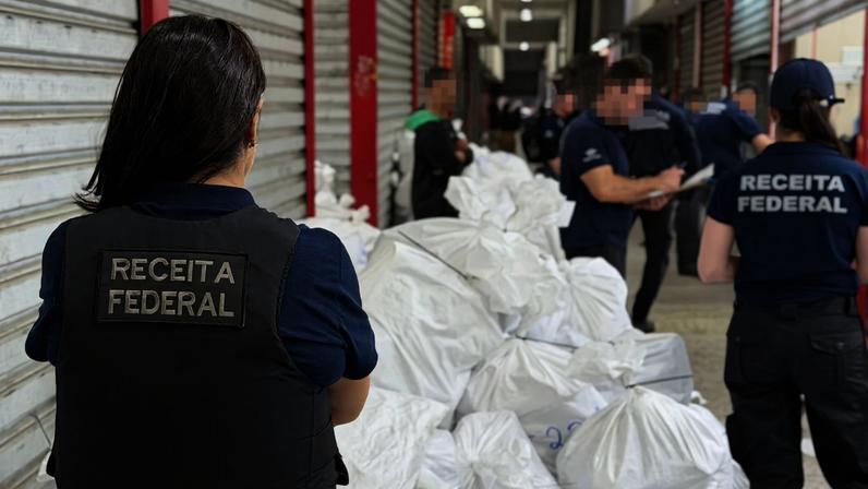 Agentes da Receita Federal em uniformes azuis e coletes à prova de balas organizam grandes sacos brancos empilhados em corredor de galpão com portas metálicas fechadas.