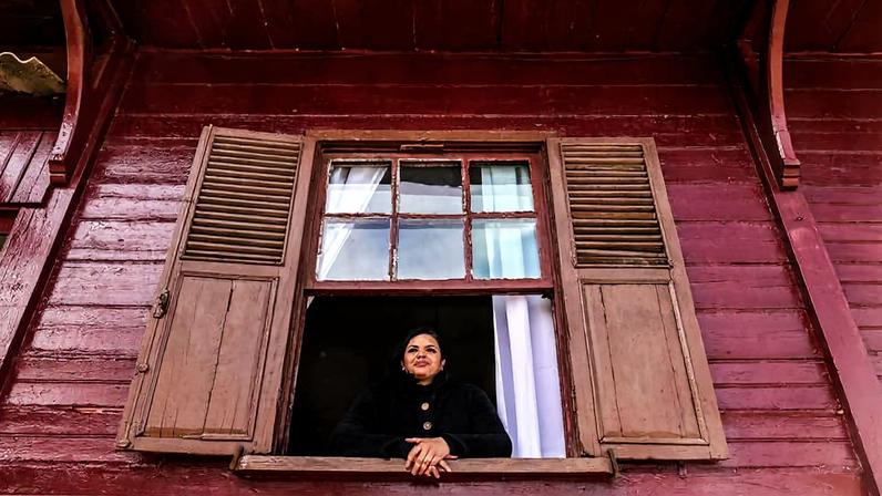 Mulher com casaco preto apoia os braços no parapeito de janela aberta em casa de madeira pintada de vermelho. Janela tem venezianas de madeira e cortina branca ao fundo.