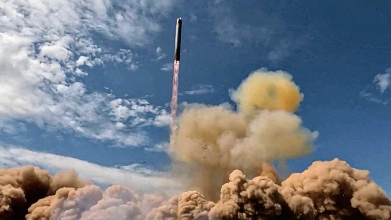 Míssil é lançado verticalmente, deixando uma trilha de fumaça rosa. Nuvens de poeira e fumaça densa se espalham na base, sob céu azul com nuvens dispersas.