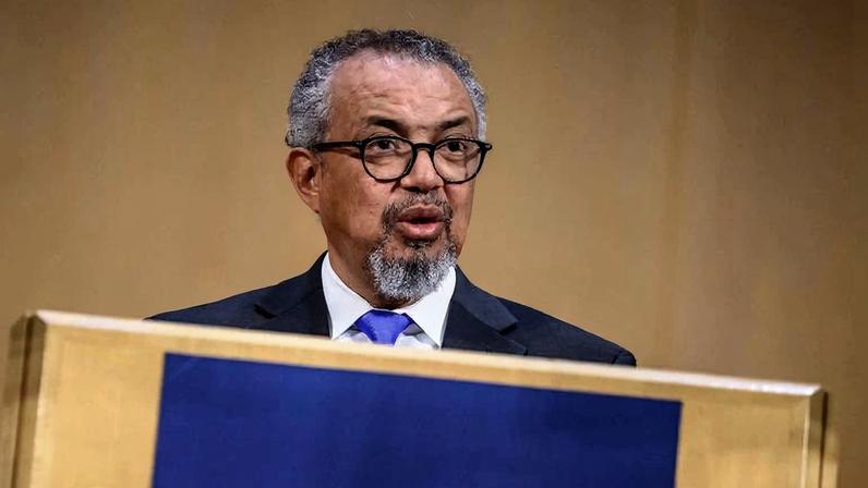 Homem de meia-idade com cabelo grisalho e barba curta usa terno escuro, camisa branca e gravata azul enquanto fala em púlpito com painel azul. Fundo neutro bege.