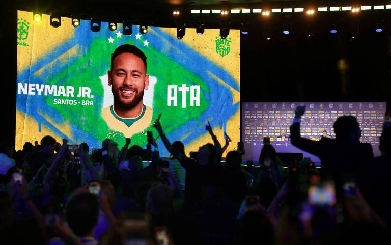 Grande tela exibe imagem de Neymar Jr. com fundo da bandeira do Brasil e texto 'NEYMAR JR. SANTOS - BRA' e 'QTA'. Público em primeiro plano levanta braços e celulares para registrar o momento em ambiente escuro com iluminação no palco.
