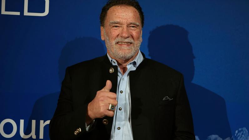 Homem de meia-idade com barba e cabelo grisalho veste camisa azul clara, blazer preto e jeans, fazendo sinal de positivo com a mão direita em frente a fundo azul com texto branco.