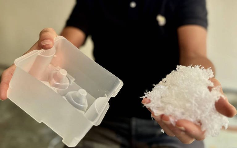 Homemcamisa preta segura em uma mão pedaços de plástico branco triturado e na outra uma caixa de plástico transparente. Bandeja plástica (à esquerda) produzida com resíduos industriais que seriam descartados
