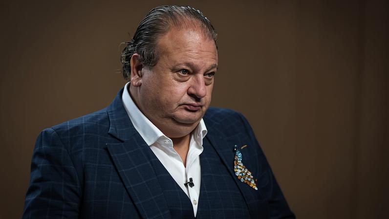 Homem de meia-idade com cabelo penteado para trás veste terno azul escuro com padrão xadrez, camisa branca e lenço colorido no bolso. Ele está em ambiente interno com fundo marrom desfocado.