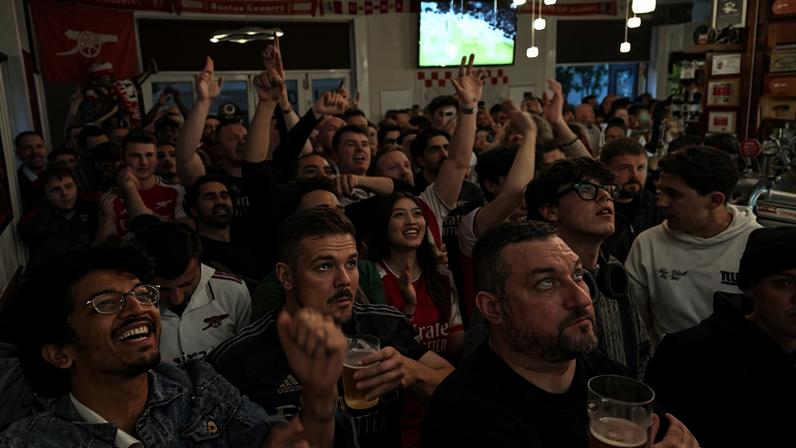 Grupo numeroso de torcedores dentro de bar, muitos vestindo camisas do Arsenal, assistindo jogo em tela grande. Alguns seguram copos de cerveja e levantam as mãos, demonstrando entusiasmo.