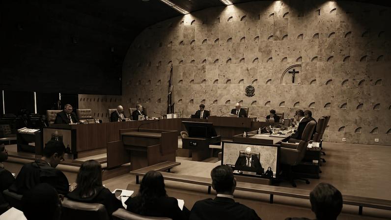 Ministros sentados em semicírculo no plenário do Supremo Tribunal Federal, com público e advogados na plateia. Parede de pedra com cruz e brasão ao fundo.