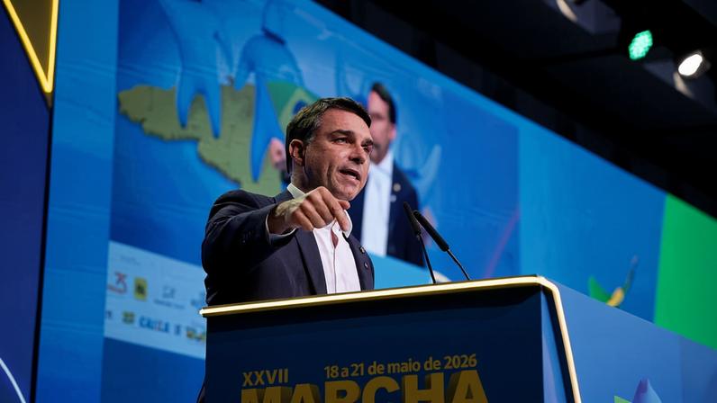 Homem de terno azul e camisa branca fala em púlpito azul com inscrições amarelas da XXVII Marcha a Brasília, realizada de 18 a 21 de maio de 2026. Fundo digital azul exibe mapa estilizado e figura desfocada.