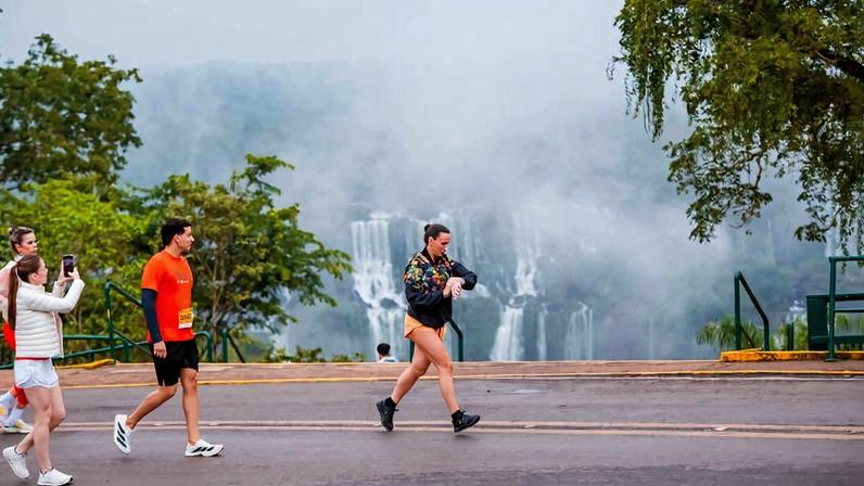 Quatro pessoas caminham em estrada asfaltada com as Cataratas do Iguaçu ao fundo, parcialmente encobertas por névoa. Vegetação verde cerca a área, e o céu está nublado.
