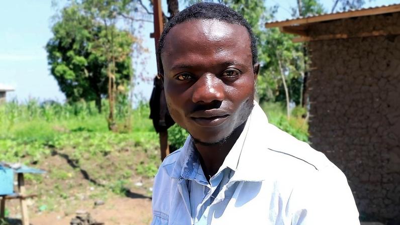 Homem negro veste camisa branca e está em área rural com vegetação e casa de barro ao fundo. Céu claro e árvores visíveis.