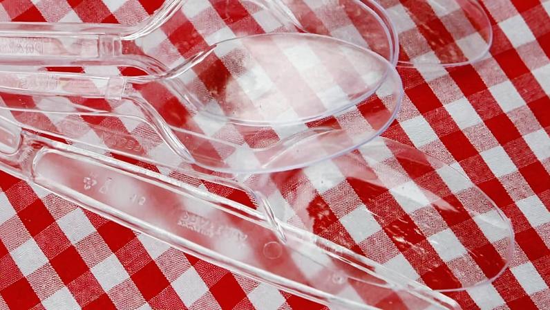 Quatro colheres plásticas transparentes dispostas sobre uma toalha de mesa com padrão xadrez vermelho e branco. As colheres estão parcialmente sobrepostas, com detalhes de reflexos visíveis na superfície lisa do plástico.