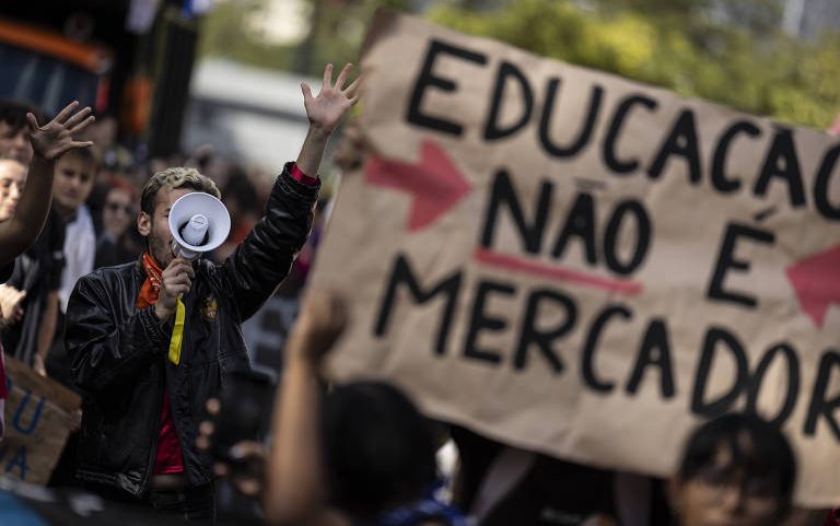 Manifestante usa megafone e levanta a mão durante protesto. Placa em destaque diz 'Educação não é mercadoria'. Pessoas ao fundo participam da manifestação em ambiente urbano.