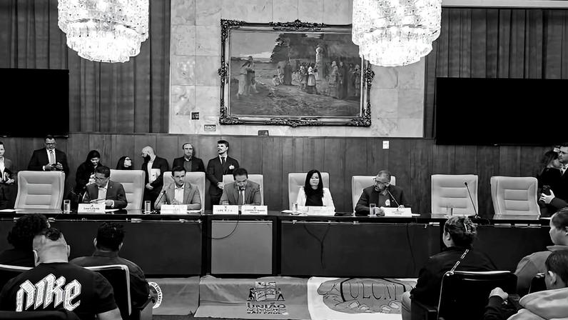 Seis autoridades sentadas em mesa com microfones em ambiente formal, atrás delas bandeira e quadro grande na parede. Público observa e fotografa a sessão.