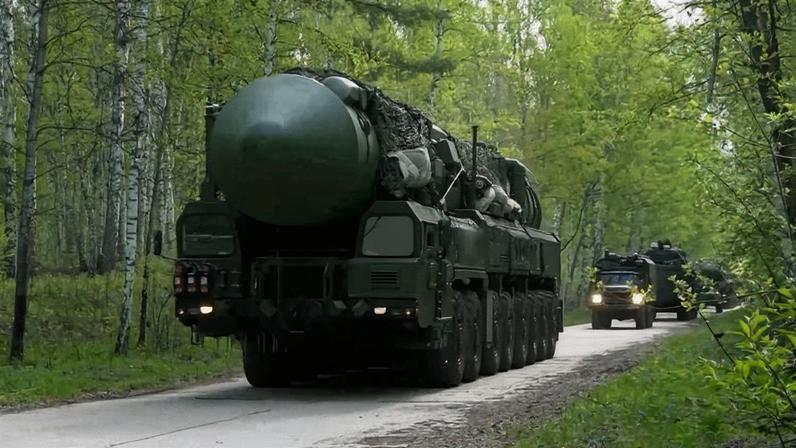 Veículo militar verde transporta míssil grande em estrada cercada por árvores verdes. Outros veículos militares seguem atrás na mesma estrada.