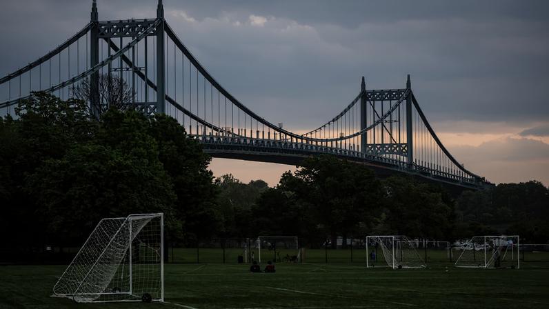 Campos de futebol vazio com várias traves brancas distribuídas sobre o gramado. Ao fundo, ponte suspensa grande com estrutura metálica cinza, cercada por árvores verdes sob céu nublado.