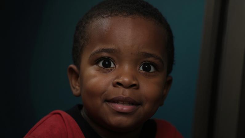Criança pequena com expressão sorridente veste camisa listrada preta e vermelha com número 10. Balões coloridos aparecem ao fundo.