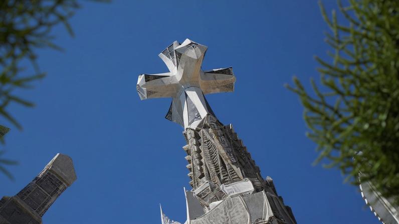 Cruz branca no topo de uma das torres da Sagrada Família em Barcelona, vista através de folhagens verdes contra céu azul claro.