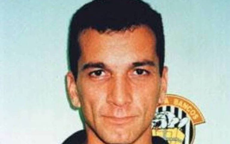 Homem de cabelo curto e escuro segura placa policial com informações sobre roubo a banco em 20 de julho de 1999, equipe Apolo 11, número de foto 1791. Fundo claro com símbolo policial parcialmente visível.