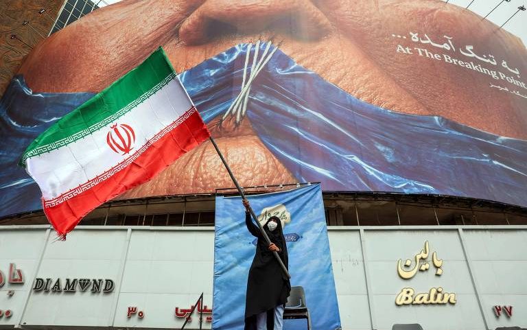 Mulher segura bandeira do Irã em frente à outdoor em Teerã que representa o presidente dos EUA, Donald Trump, e o fechamento do estreito de Hormuz