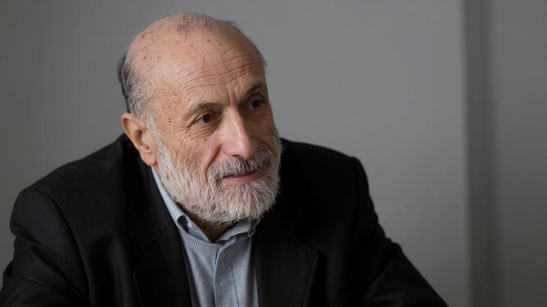 Homem idoso com barba branca e cabelo ralo veste blazer escuro sobre suéter claro, olhando levemente para a esquerda em fundo neutro.