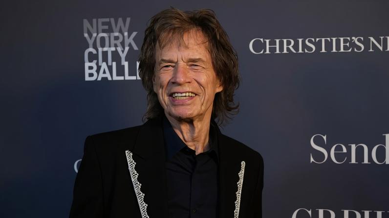 Homem de cabelos castanhos claros e ondulados veste paletó preto com detalhes brancos e camisa azul escura, posando em frente a painel cinza com textos do New York City Ballet e Christie's.