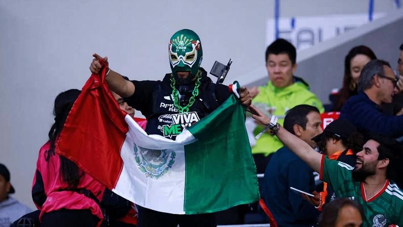 Torcedor com máscara verde e corrente no pescoço segura bandeira do México em arena esportiva. Pessoas ao redor assistem e interagem, algumas vestindo camisas verdes da seleção mexicana.