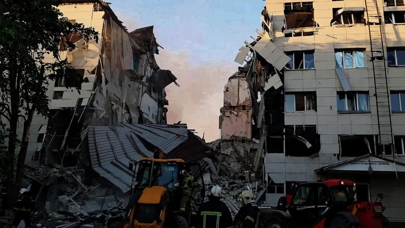 Prédio de vários andares com fachadas destruídas e escombros no chão entre duas estruturas danificadas. Dois veículos de construção e três trabalhadores com capacetes e coletes refletores estão na área de resgate.