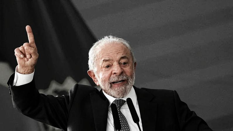 Homem de cabelos brancos e barba grisalha, vestido com terno azul escuro e gravata, fala gesticulando com a mão direita levantada. Ao fundo, bandeira do Brasil desfocada.