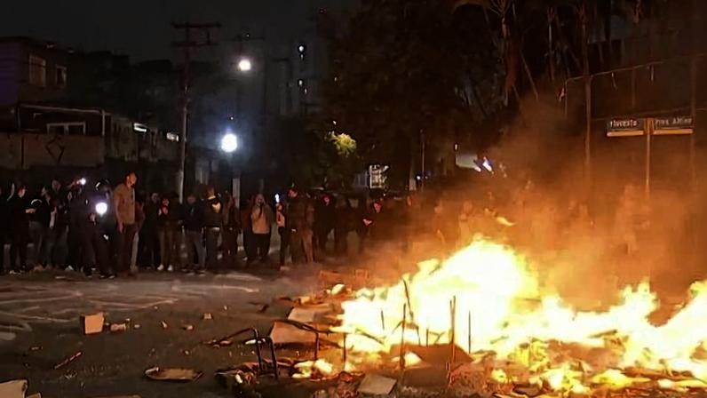 Grupo de pessoas reunidas à noite em rua bloqueada por fogo e objetos queimando. Luzes de postes iluminam a cena e cones de trânsito estão no chão.